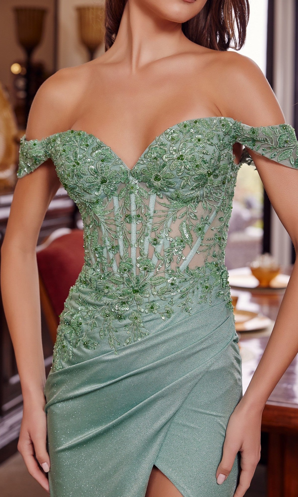 A woman stands indoors wearing the Nox Anabel H1879, a long green prom dress with beaded floral embroidery, strapless off-the-shoulder design, and sheer bodice.